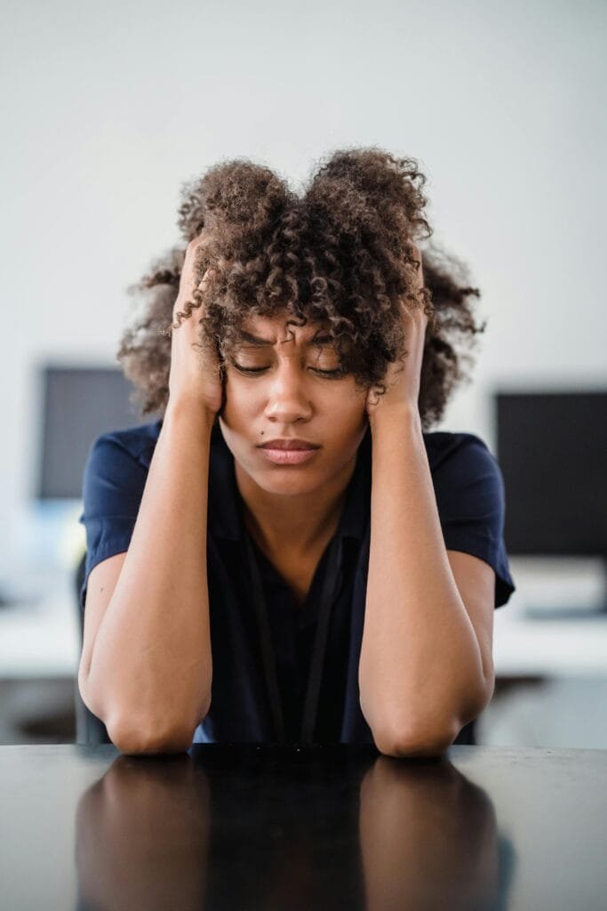 Woman in office setting expresses stress, seated at desk with hands in hair. Perfect for workplace stress concept.